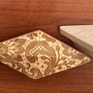 Könnte beinhalten: Zwei dekorative Holzstücke. Eines ist ein helles, rautenförmiges Objekt mit einem komplizierten, geätzten Blumendesign. Das andere ist ein schlichtes, rautenförmiges Holzstück. Beide liegen auf einer braunen Holzoberfläche.