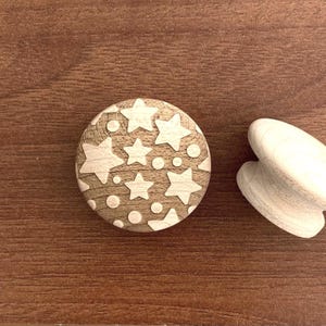 Könnte beinhalten: Ein hölzerner Schrankknopf mit einem Stern- und Punktmuster. Der runde Knopf hat eine helle Holzoberfläche mit weißen Stern- und Punktausschnitten. Ein zweites, helles Holzobjekt befindet sich neben dem Knopf.
