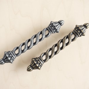 Può includere: Due maniglie per mobili in metallo ornate con un design a torsione e un terminale a forma di corona. Una maniglia è argentata e l'altra è dorata.