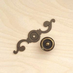 May include: A bronze-toned metal drawer pull consisting of a circular knob with a textured centre and a separate, elongated, decorative backplate featuring curved, scroll-like ends and a central circular opening for mounting hardware.