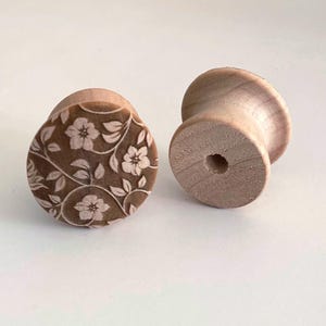 May include: Two light-coloured wooden spools. One spool features a dark brown circular top surface engraved with a detailed floral pattern of five-petaled flowers and leaves. The second spool is plain, showing the natural wood grain and a centre hole.