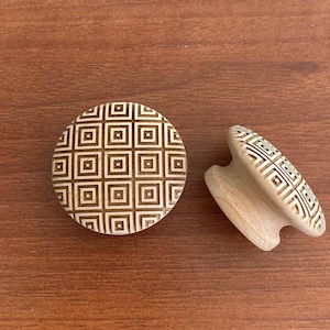 Peut inclure: Deux boutons de meuble en bois avec un motif géométrique carré dans un coloris marron clair et crème. L'un des boutons est rond, l'autre est en forme de poignée. Les boutons sont sur une surface en bois marron.