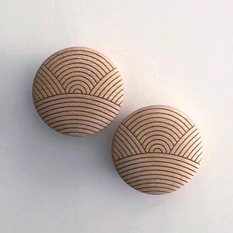 May include: Two round wooden cabinet knobs with a light brown finish. Each knob features a carved design of curved lines, resembling a stylized landscape or sunrise. The knobs are set against a white background.