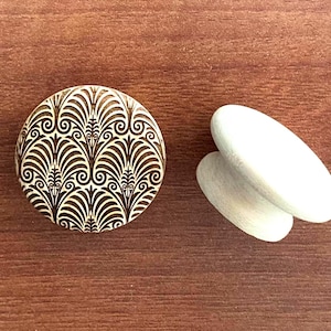 Peut inclure: Un bouton de meuble rond en bois avec un motif Art déco brun et crème. Deux tirettes de tiroir ovales en bois blanc sont à côté du bouton. Les objets sont sur une surface en bois brun.
