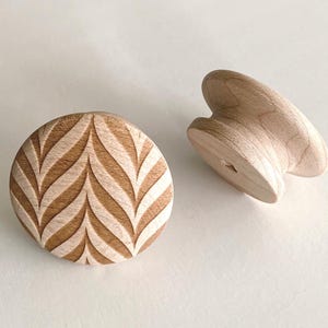 May include: Two unfinished wooden cabinet knobs. One is round with a carved leaf pattern, and the other is a mushroom-shaped knob. The knobs are light brown and appear to be made of natural wood.