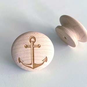 Puede incluir: Dos pomos de armario de madera. Uno es redondo con un diseño de ancla grabado. El otro es un pomo de madera a juego con una forma curva. Ambos son de madera de color claro, probablemente arce o abedul.