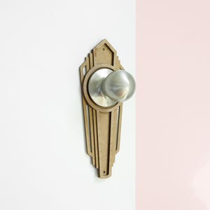 Peut inclure: Un cache-bouton de porte en bois avec un design Art Déco. Le cache est de couleur brun clair et a un bouton de porte argenté au centre.