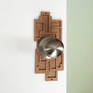 Könnte beinhalten: Ein dekorativer Türknauf mit geometrischem Design. Der Knauf besteht aus gebürstetem Metall und ist auf einer braunen, lasergeschnittenen Holzrückplatte angebracht. Die Rückplatte ist an einer weißen Wand montiert.