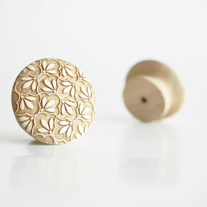 Op de afbeelding: Twee houten kastknoppen. Eén is scherp, met een beige bloemenpatroon op een rond oppervlak. De andere knop is onscherp. De knoppen zijn gemaakt van hout en staan tegen een witte achtergrond.