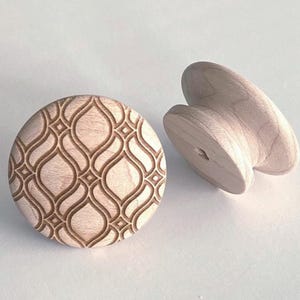 Op de afbeelding: Twee onafgewerkte houten kastknoppen. Eén is rond met een uitgesneden, zich herhalend geometrisch patroon. De andere heeft een meer traditionele vorm met een gebogen handvat. Beide knoppen zijn lichtgekleurd hout.