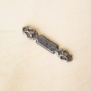 May include: A silver-toned metal drawer pull with a decorative, ornate design. The pull has a single hole for mounting.
