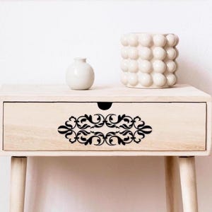 May include: A light-colored wooden nightstand with a drawer featuring a black decorative design. A small white vase and a textured white ceramic pot sit on top. The nightstand has four tapered legs.