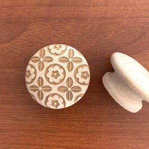 May include: A wooden stamp with a floral pattern and a wooden massage tool. The stamp is round with a repeating flower design. The massage tool is heart-shaped and made of light-coloured wood. Both items are on a brown surface.