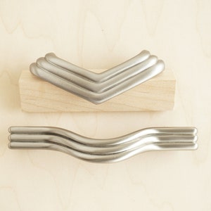 May include: Two silver cabinet pulls, one shaped like a chevron and the other shaped like a wave. Both pulls have a triple-layered design.