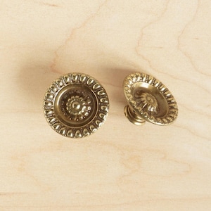 May include: Two gold-toned metal cabinet knobs with a decorative, ornate design. The knobs have a raised, circular centre with a floral pattern.