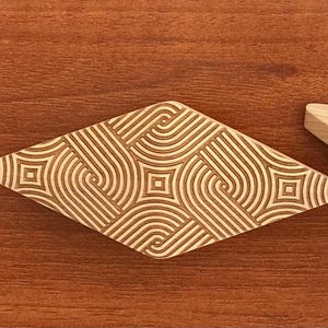 Könnte beinhalten: Dekoratives Objekt aus Holz in Diamantenform mit einem geometrischen Muster aus geschwungenen Linien und Quadraten. Das Objekt ist hellbraun mit einem dunkleren braunen eingravierten Design. Ein zweites, kleineres Holzobjekt ist sichtbar.