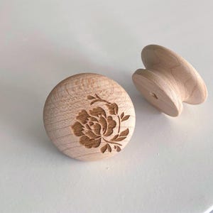 Op de afbeelding: Twee onafgewerkte houten kastknoppen. Eén is rond met een bloemenontwerp in het hout geëtst. De andere is een knop in de vorm van een handvat. Beide zijn gemaakt van lichtgekleurd hout.