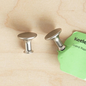 May include: Two silver cabinet knobs with a round top and a short, cylindrical base. The knobs are on a light wood surface.
