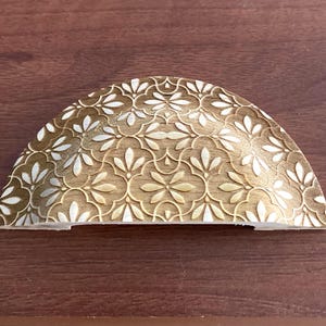 Könnte beinhalten: Ein dekorativer, halbmondförmiger Schrankgriff mit floralem Muster. Die Hardware hat eine warme Messingfarbe mit weißen Akzenten, vor einem dunklen Holzuntergrund. Das Design zeigt ein sich wiederholendes Blumenmotiv.