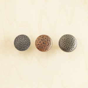 Op de afbeelding: Drie ronde metalen kastknoppen in verschillende afwerkingen: zilver, koper en zwart. Elke knop heeft een gehamerde textuur.