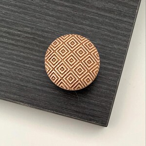 Peut inclure: Un bouton de meuble rond en bois avec un motif géométrique en losanges. Le bouton est de couleur marron clair et repose sur une surface gris foncé. Le motif est une répétition de carrés dans des carrés.