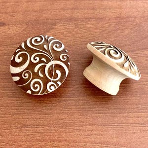 May include: Two wooden ear plugs with a decorative swirl design. One plug is flat and round, while the other is curved. The plugs are light-coloured wood with a dark brown swirl pattern. The background is a wooden surface.