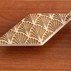 Könnte beinhalten: Ein helles Holzschubladengriff mit einem fächerförmigen Design. Das dekorative Element ist in das Holz geschnitzt und erzeugt eine strukturierte Oberfläche. Der Schubladengriff ist rautenförmig.