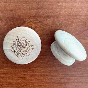 May include: A wooden disc with a rose engraving and two oval wooden objects. The rose design is etched into the wood. The objects are light-coloured and sit on a brown wooden surface.