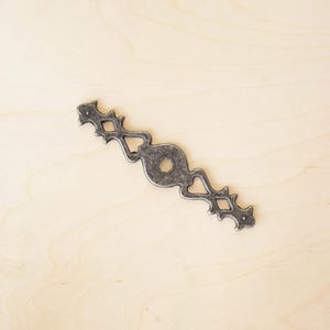 Könnte beinhalten: Ein silberfarbener Metall-Schubladengriff mit einem dekorativen Design, das ein zentrales Oval mit einem Loch und komplizierten floralen Mustern auf beiden Seiten aufweist.