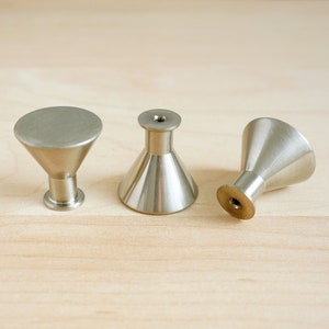 Op de afbeelding: Drie zilveren metalen kastknoppen met een kegelvorm. De knoppen hebben een gladde, gepolijste afwerking.