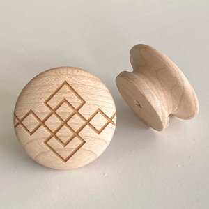 Op de afbeelding: Twee onafgewerkte houten kastknoppen. Eén knop is rond met een gegraveerd geometrisch ontwerp. De andere knop heeft de vorm van een paddenstoel. Beide zijn gemaakt van lichtgekleurd hout en staan tegen een witte achtergrond.