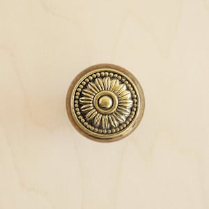 Op de afbeelding: Een ronde, messingkleurige kastknop met een decoratief bloemmotief. De knop heeft een verhoogd, rond midden en een parelrand.