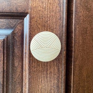 May include: A round, light-colored wooden cabinet knob with a carved landscape design. The knob is mounted on a dark brown wooden cabinet door. The design features horizontal and curved lines.