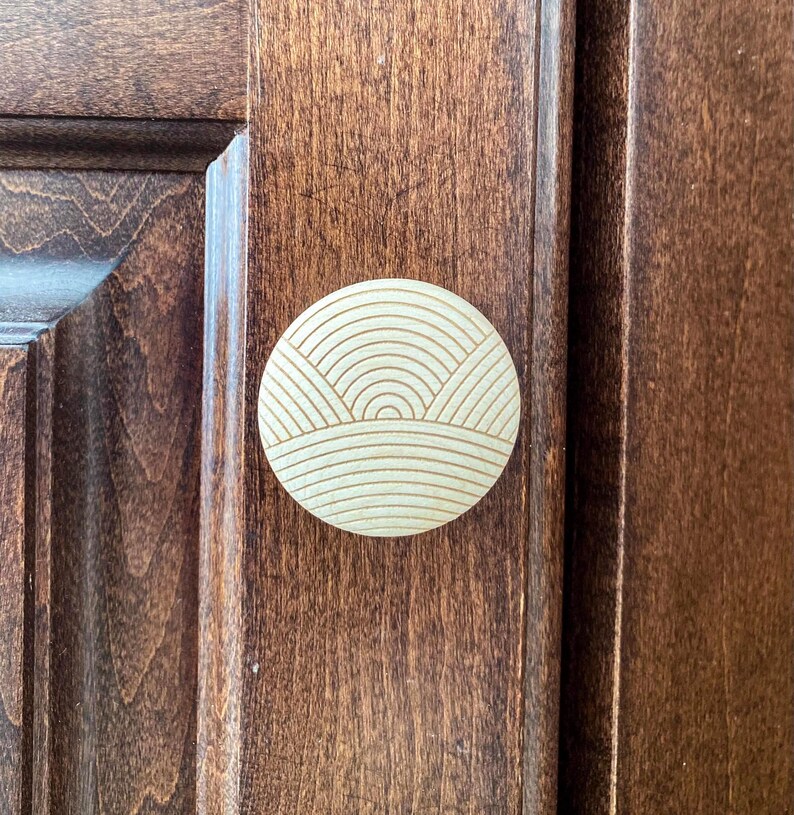 May include: A round, light-colored wooden cabinet knob with a carved landscape design. The knob is mounted on a dark brown wooden cabinet door. The design features horizontal and curved lines.