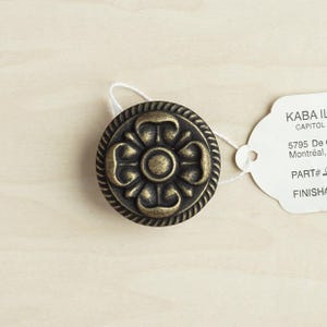 Könnte beinhalten: Ein runder, verzierter, messingfarbener Schrank-Knopf mit einem floralen Design. Der Knopf hat eine dunkelbraune Oberfläche und eine erhabene, kreisförmige Mitte.