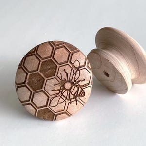 Puede incluir: Dos pomos de madera para armarios. Uno presenta un patrón de panal con una abeja grabada en el centro. El otro es un diseño de madera maciza sin terminar. Los pomos son de color marrón claro y están sobre una superficie blanca.