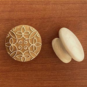 May include: A wooden, round decorative piece with an engraved floral design, next to two smooth, oval-shaped wooden objects. The items are light brown and sit on a darker brown wooden surface.