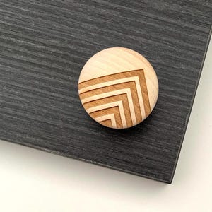 May include: A circular wooden knob in a light colour, featuring a geometric chevron pattern. The knob is set against a dark grey, textured surface. The chevron design is created by etched lines, adding a tactile element to the design.