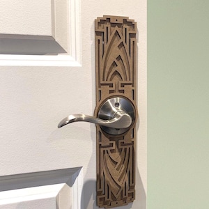 May include: A door handle with a decorative wooden backplate featuring an Art Deco design. The handle is made of brushed metal and is mounted on a white door. The backplate is a light brown colour.