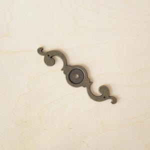 Peut inclure: Une poignée décorative en métal pour armoire avec un motif enroulé et un centre circulaire. La poignée est de couleur or clair.