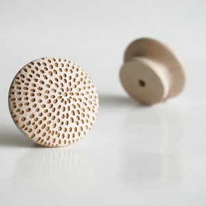 May include: Two unfinished wooden cabinet knobs. One knob is in focus, showing a circular design with many small, evenly spaced holes. The other knob is slightly out of focus. The knobs are light beige in color and appear to be made of wood.