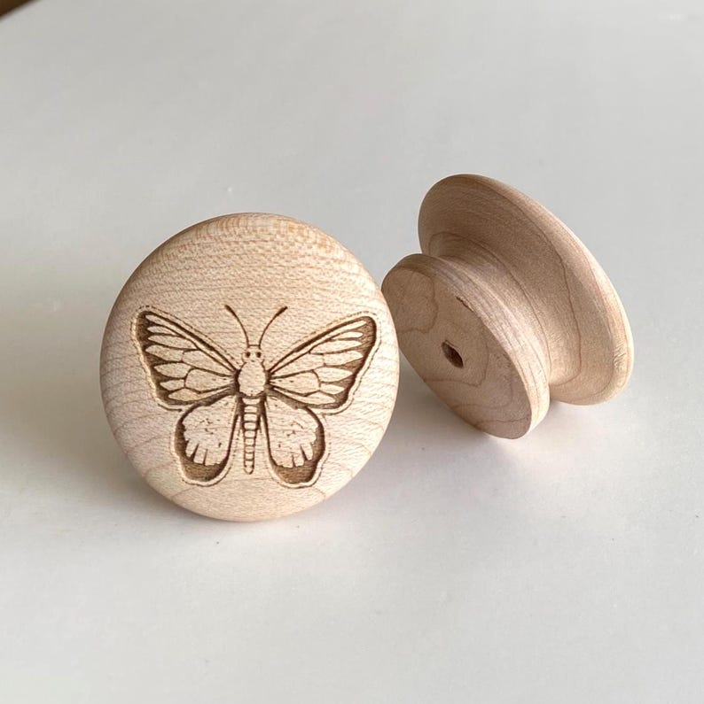 May include: Two unfinished wooden cabinet knobs. One knob features a detailed butterfly engraving. The other knob has a rounded shape with a hole for mounting. The knobs are made of light-coloured wood.