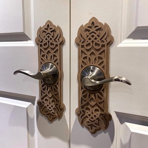 May include: Two wooden door handle covers with detailed, symmetrical cut-out patterns. The covers are light brown and are mounted on white doors. The handles are silver-toned, creating an ornate and decorative appearance for interior design.