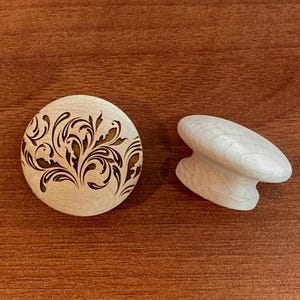 May include: Two wooden cabinet hardware pieces. One is a round knob with a dark brown floral design. The other is a white, curved handle. Both are on a brown wooden surface.