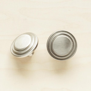 Może przedstawiać: Dwa srebrne, okrągłe gałki do szafki z wzorem koncentrycznych kół. Gałki mają szczotkowane wykończenie.