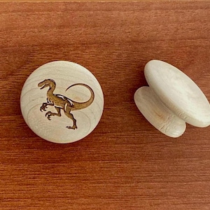 以下が含まれることがあります： レーザー彫刻された恐竜のデザインが施された木製の丸いディスクと、2つの滑らかな楕円形の木片。 恐竜は走っているポーズで描かれています。 木目調の背景に配置されています。