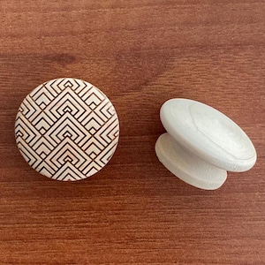Könnte beinhalten: Zwei Holzschrankknöpfe. Einer ist rund mit einem geometrischen Muster in Braun und Beige. Der andere ist ein Paar oval geformter Knöpfe in hellem Holz. Die Knöpfe liegen auf einer braunen Holzoberfläche.