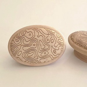 May include: Two oval-shaped wooden knobs featuring a light beige natural wood grain finish. The front surface of each knob is engraved with a dark brown, intricate topographic map pattern consisting of continuous, flowing, irregular contour lines.