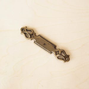 May include: A gold-toned metal drawer pull with an ornate design. The pull has a rectangular centre plate with a single hole for mounting.
