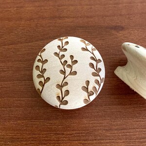 May include: A light-colored wooden cabinet knob with a floral design and a matching round wooden cabinet knob with a similar floral design. The knobs are on a brown wooden surface.